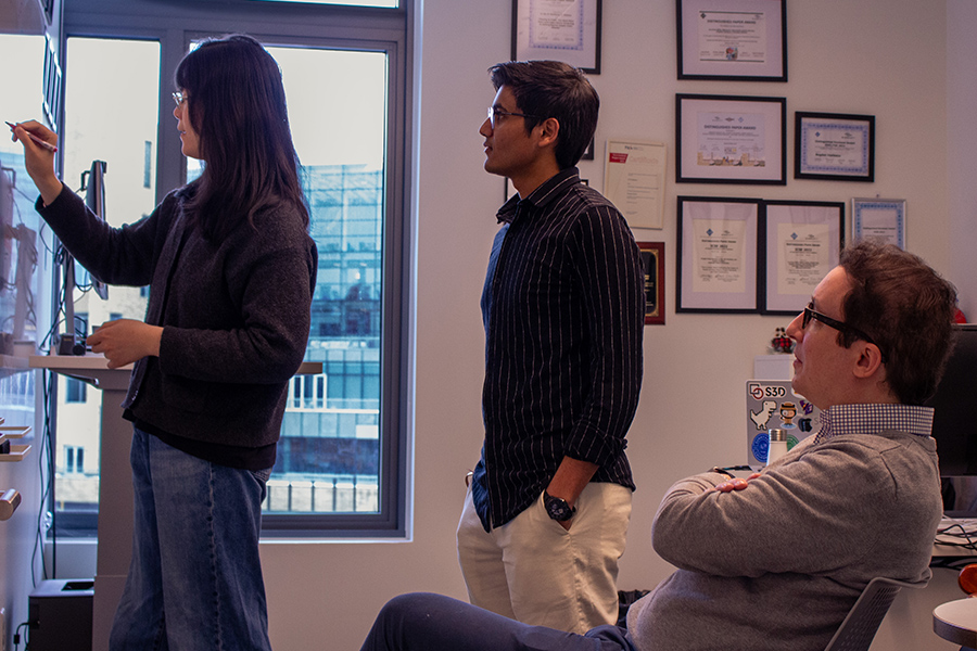 Two people stand in front of a white board, one writing and one watching, while a seated man looks on.